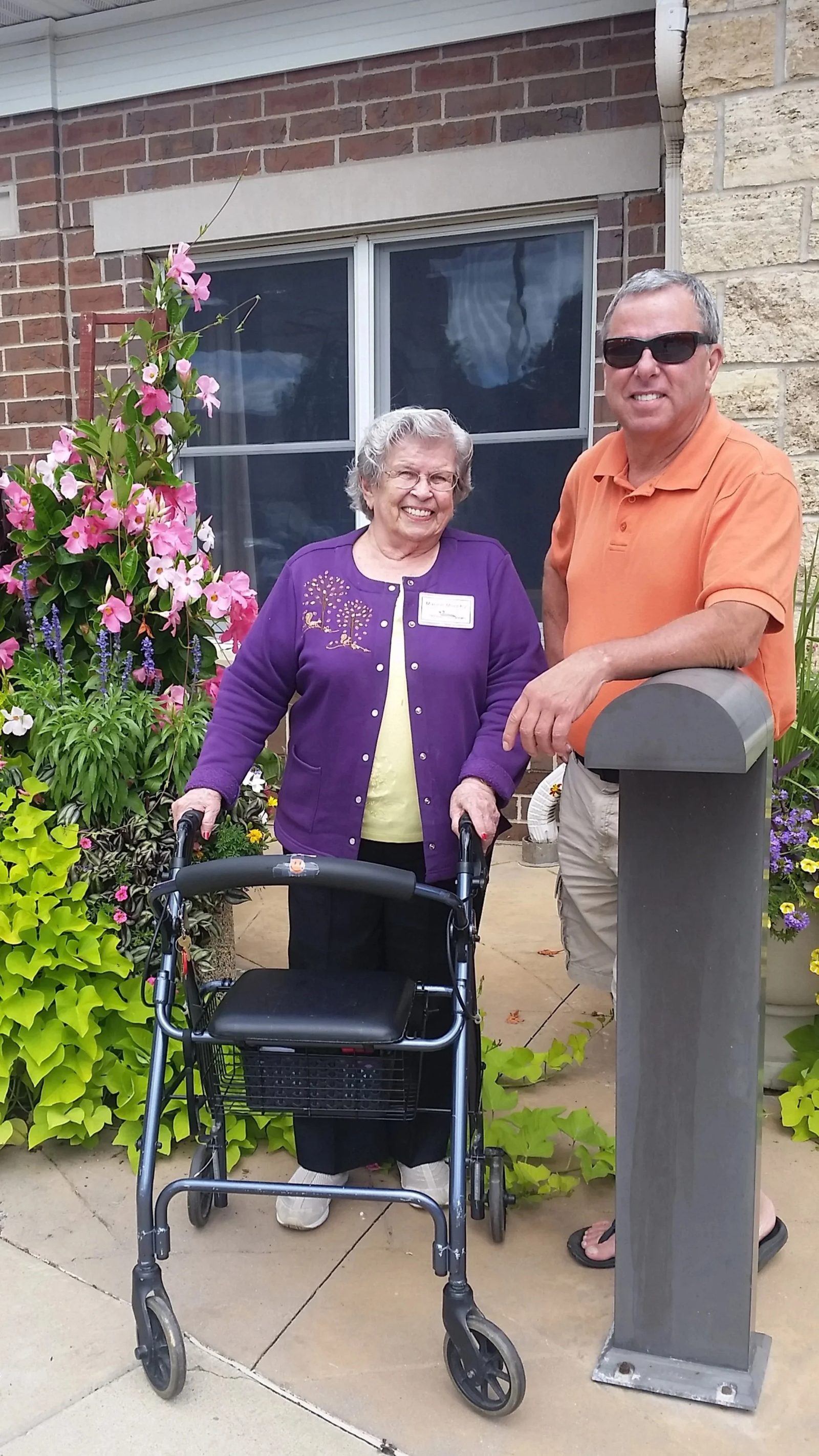 Kevin with an older woman and her walker outside a flower-lined building